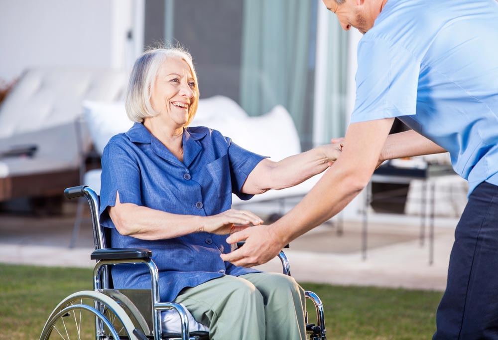 Caretaker Helping Senior Woman To Get Up From Wheelchair